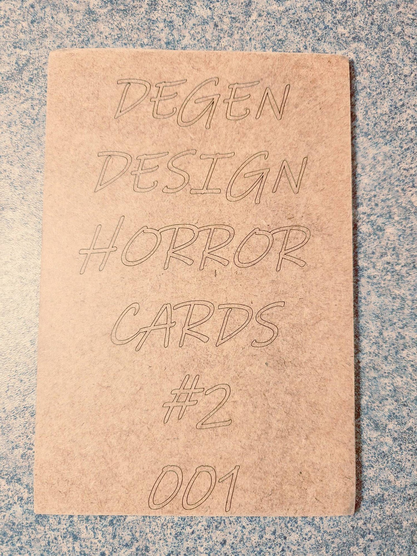 Back of Degen Design Serial Killer Collector Card #2 Ted Bundy with engraved text 'Degen Design Horror Cards #2 001' on textured surface.